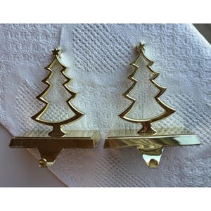 May include: A pair of gold-toned metal stocking holders, each featuring a cutout silhouette of a Christmas tree topped with a small star. The base of each holder is rectangular and includes a downward-facing hook for hanging stockings.