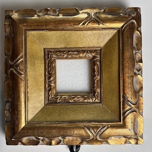 May include: Square, ornate picture frame with a gold finish. The frame has a layered design with a smaller, inner frame. The outer frame has a textured, carved pattern. The inner frame is a lighter gold color. A black ribbon is attached to the bottom.