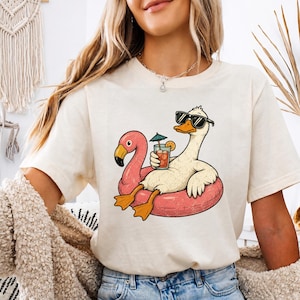Könnte beinhalten: Cremefarbenes T-Shirt mit einer Cartoon-Gans, die eine Sonnenbrille trägt und einen Cocktail hält, während sie auf einem rosa Flamingo-Schwimmer treibt. Die Gans hat orangefarbene Füße und einen gelben Schnabel. Das Shirt hat eine lockere Passform.