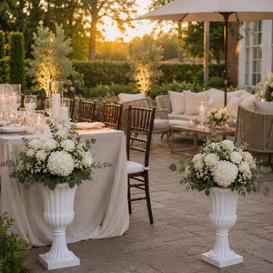 May include: Two white fluted pedestal urns, each holding a large, rounded floral arrangement of white hydrangeas, white roses, and green eucalyptus foliage. The urns feature a classic flared rim and a square base, standing on a stone patio.