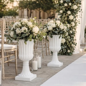May include: Two white fluted pedestal urns holding large arrangements of white roses and green eucalyptus leaves. The urns feature a classic flared rim and a square base, positioned on a stone floor alongside gold chairs and glass candle holders.