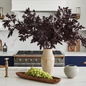 May include: A modern kitchen with navy blue cabinets, a white countertop, and gold fixtures. A wooden tray holds green grapes. A beige vase with dark purple branches sits near the stove. Two pendant lights hang above the island.