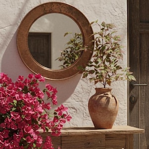 May include: An outdoor scene featuring a wooden console table with a terracotta vase holding greenery. A round copper mirror hangs on the textured white wall. A large wooden door and a terracotta pot with pink flowers complete the scene. A candle lantern sits on the table.