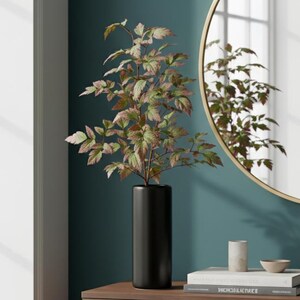 May include: A black cylindrical vase holds a leafy plant with green and reddish-brown leaves. The vase sits on a wooden cabinet, next to a stack of books and a small bowl. A round gold-framed mirror hangs on the teal wall.