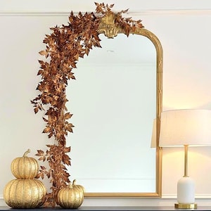 May include: A navy blue sideboard with gold hardware, topped with gold pumpkins and a lamp. A gold-framed mirror and fall foliage garland hang above. Woven baskets sit below, creating a stylish, seasonal display.