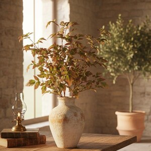 May include: A rustic interior scene featuring a wooden table with a distressed white vase holding leafy branches. A small oil lamp and stacked books sit on the table. An olive tree in a terracotta pot and a beige sofa are in the background.