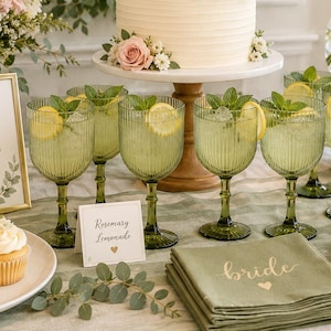 May include: A two-tier white cake decorated with pink roses on a wooden stand, surrounded by green ribbed glasses of lemonade with lemon slices, cupcakes, macarons, and floral arrangements with signs reading welcome to the bridal shower and sip sip hooray.