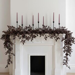 May include: A white fireplace mantel decorated with a dark burgundy garland and six mauve taper candles in black holders. The garland has dark leaves and is draped across the mantel.