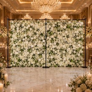 May include: A large rectangular backdrop covered in dense white flowers, including roses and hydrangeas, supported by a black metal frame. The floral wall stands in a grand room with marble floors and crystal chandeliers hanging from the ceiling.
