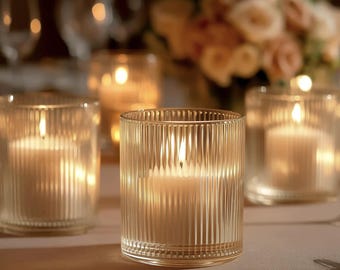 May include: Several lit candles in clear, ribbed glass holders. The candles are white and the flames are bright. The background includes a floral arrangement and other candles, creating a warm, inviting ambiance.
