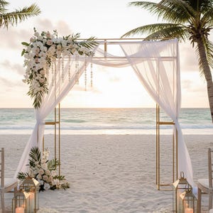 May include: A gold metal wedding arch on a sandy beach, decorated with white fabric, flowers, and greenery. The arch is set against the ocean with palm trees and lanterns with candles.