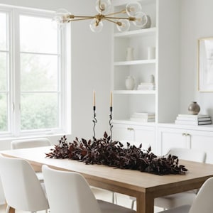 May include: A dining room scene featuring a light wood table with white chairs. A dark burgundy floral arrangement runs down the center of the table, with two black candle holders and candles. A gold chandelier hangs above.