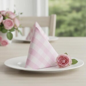 May include: A white plate with a pink and white checkered napkin folded into a triangle. A pink rose with green leaves rests on the plate. A vase of pink roses is in the background.
