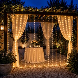 May include: A wooden pergola structure draped with sheer white curtains and vertical strings of warm white fairy lights. A round table with a white tablecloth and a floral centerpiece sits in the center, illuminated by the glowing light strands.