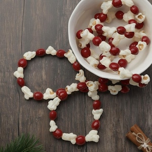 May include: A close-up of popcorn and red candy garland, with some in a white bowl. The garland is arranged on a dark wood surface, with a sprig of evergreen and cinnamon sticks in the background.