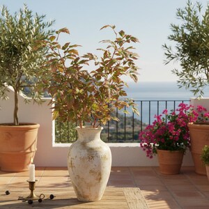 May include: A large, off-white ceramic vase with a distressed finish holds leafy branches with green and reddish-brown leaves. The vase sits on a wooden table with a lit candle in a brass holder. Olive trees and pink flowering plants are in terracotta pots.