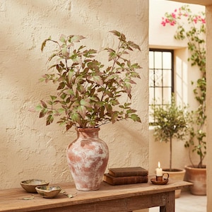 May include: A rustic, terracotta vase filled with leafy branches, displaying green and reddish-brown foliage. The vase sits on a wooden table with small bowls and books. A lit candle and outdoor plants are visible in the background.