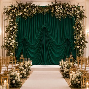 May include: A deep emerald green fabric backdrop draped in a gathered style, framed by an arch of white roses and greenery. The scene includes a white aisle runner, gold metal lanterns with lit candles, and floral arrangements on the floor.