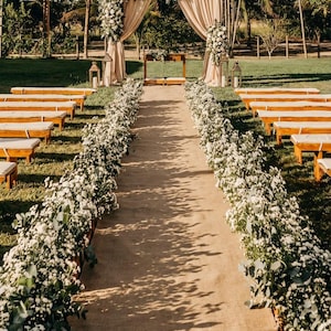 May include: Outdoor wedding ceremony setup with a pathway lined with white flowers. A floral arch and draped fabric create a focal point. Wooden benches are on the sides, and palm trees are in the background.
