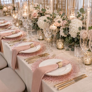 May include: A formal table setting featuring white ceramic plates, blush pink fabric napkins secured with gold metal rings, textured rose gold charger plates, gold-toned flatware, clear crystal glassware, and lit candles in glass cylinders.