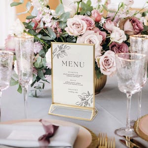 May include: A gold-framed menu card with the word "MENU" in a serif font, displayed on a table setting. The menu lists starter, main, and dessert options. The background features a floral arrangement with pink and white roses.
