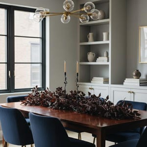 May include: A dining room set with a dark wood table, navy blue chairs, and a gold chandelier. A burgundy leaf garland runs down the center of the table. Built-in white shelves and a black window frame complete the room's decor.