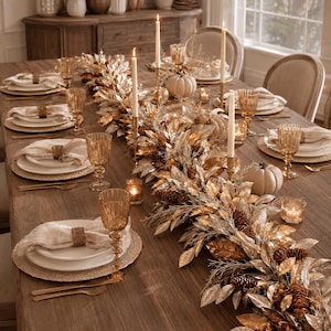 May include: A dining table set for a meal featuring white plates, textured placemats, amber-colored glasses, and gold-toned napkin rings. A long centerpiece runs down the middle, composed of metallic gold and silver leaves, pinecones, and candles.