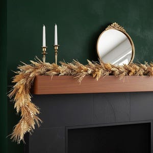 May include: A decorative mantel arrangement featuring a dried wheat garland, two white candles in gold candlesticks, and a gold-framed oval mirror. The arrangement is set against a dark green wall and a black fireplace.