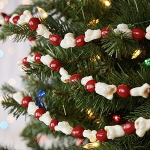 May include: A Christmas tree decorated with a garland of red beads and white popcorn. The garland is strung across the green branches, with colorful lights in the background. A festive holiday decoration.