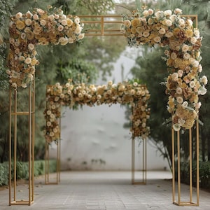 May include: A golden metal wedding arch adorned with cream and tan flowers. The arch is rectangular with floral arrangements at the corners and along the top. The scene is set outdoors with greenery in the background.