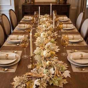 May include: A long wooden dining table set for a meal with white plates, beige napkins with gold rings, amber-colored wine glasses, and tall white taper candles in brass holders. A centerpiece of faux autumn leaves and small white pumpkins runs down the center.