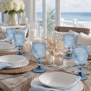 May include: A set of light blue pressed glass goblets featuring a detailed embossed pattern on the bowl and a sturdy stem. The goblets are arranged on a dining table alongside white ceramic bowls, woven placemats, and coastal decor.