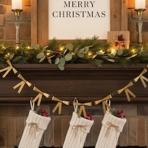 May include: A Christmas mantel decorated with a wooden sign that reads "MERRY CHRISTMAS". The mantel is adorned with greenery, lights, and three cream-colored knit stockings filled with berries. Two lit candles flank the sign.