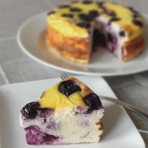 May include: A slice of blueberry cheesecake on a white square plate with a fork. The cake has a yellow top with blueberries and a creamy white interior with purple blueberry swirls. Another cheesecake is in the background.