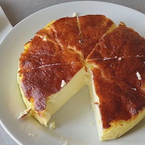 May include: A partially sliced Basque burnt cheesecake on a white plate, with a separate plate holding a single slice. The cheesecake has a dark, caramelized exterior and a creamy, pale interior. A fork rests beside the slice.