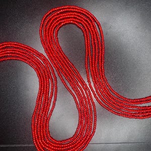 Könnte beinhalten: Leuchtend rote Bauchkette, bestehend aus kleinen, runden Perlen, die zusammen aufgefädelt sind. Die Perlen sind in mehreren Strängen angeordnet und bilden ein fließendes, elegantes Design. Die Perlen werden vor einem dunklen Hintergrund gezeigt.