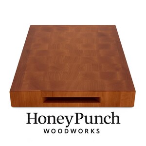 May include: A rectangular, end-grain wooden cutting board with a warm brown hue. The board features a built-in handle and the text "HoneyPunch WOODWORKS" in black. The wood grain pattern is visible across the surface.