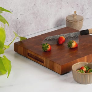 May include: A rectangular, end-grain wooden cutting board with a handle cutout. A Japanese-style knife rests on the board, next to three strawberries. A small ceramic bowl with strawberries and a small ceramic jar are also present.