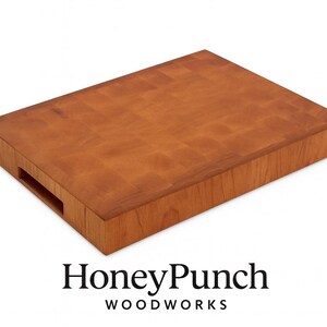 May include: A rectangular, end-grain cutting board made of light brown wood. The board has a handle cutout on one side and the HoneyPunch Woodworks logo at the bottom. The wood grain pattern is visible on the surface.