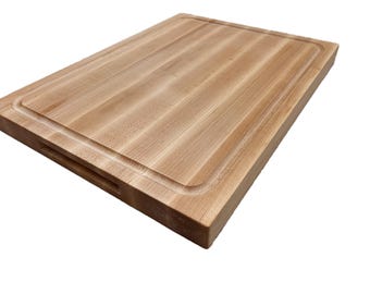 May include: A rectangular wooden cutting board with a juice groove around the edge. The board is made of light-colored wood and has a handle cut out on one side. The cutting board is designed for food preparation.
