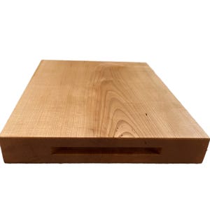 May include: A rectangular wooden cutting board with a light brown finish. The board has a recessed handle on one side. The wood grain is visible across the surface. This kitchen item is ideal for food preparation.