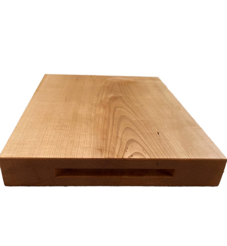 May include: A rectangular wooden cutting board with a light brown finish. The board has a recessed handle on one side. The wood grain is visible across the surface. This kitchen item is ideal for food preparation.