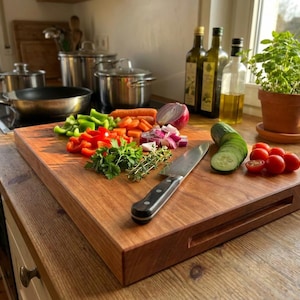 Pode incluir: Uma tábua de corte de madeira com uma variedade de vegetais frescos, incluindo pimentões vermelhos e verdes, cenouras e pepinos. Uma faca de aço inoxidável repousa na tábua, junto com ervas e tomates. Garrafas de azeite no fundo.