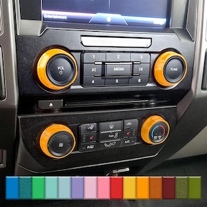 May include: A car dashboard featuring orange trim rings around the radio and climate control knobs. The dashboard includes buttons for media, volume, and climate control. A colour palette of various colours is shown at the bottom.
