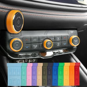 Puede incluir: Interior de coche con mandos de control naranjas y negros y una selección de llaveros de silicona de colores. Los mandos incluyen controles de volumen y pantalla. Los llaveros son de varios colores, incluyendo azul, verde, rosa y naranja.