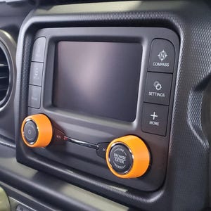 May include: Close-up of a car's dashboard with a central display screen. Orange knobs and buttons are visible, including "Compass," "Settings," and "More." The screen is black, and the dashboard has a textured, dark gray finish.
