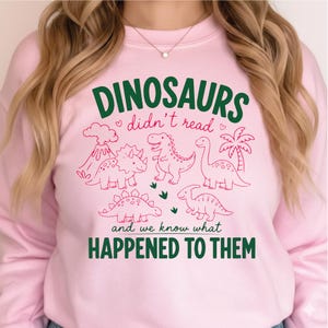 May include: A light pink sweatshirt featuring a dark green and red graphic print. The text reads DINOSAURS didn't read and we know what HAPPENED TO THEM. The design includes line art illustrations of four dinosaurs, a volcano, and a palm tree.