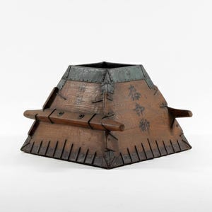 May include: A wooden, pyramid-shaped container with metal trim and handles. The container has a dark brown finish with visible wood grain and is adorned with Japanese characters. The container is a unique decorative item.