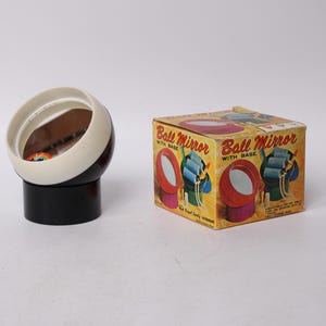 May include: A vintage ball mirror with a base, featuring a white and brown sphere with a reflective surface. The mirror sits on a black cylindrical base. A colorful box with the text "Ball Mirror with Base" is also present.