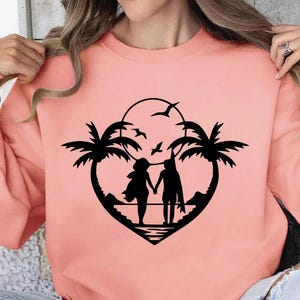 May include: A coral-colored sweatshirt featuring a black silhouette design. The design depicts a couple holding hands on a beach, framed by palm trees and a heart shape. Birds fly in the sky above the couple, with a sun in the background.